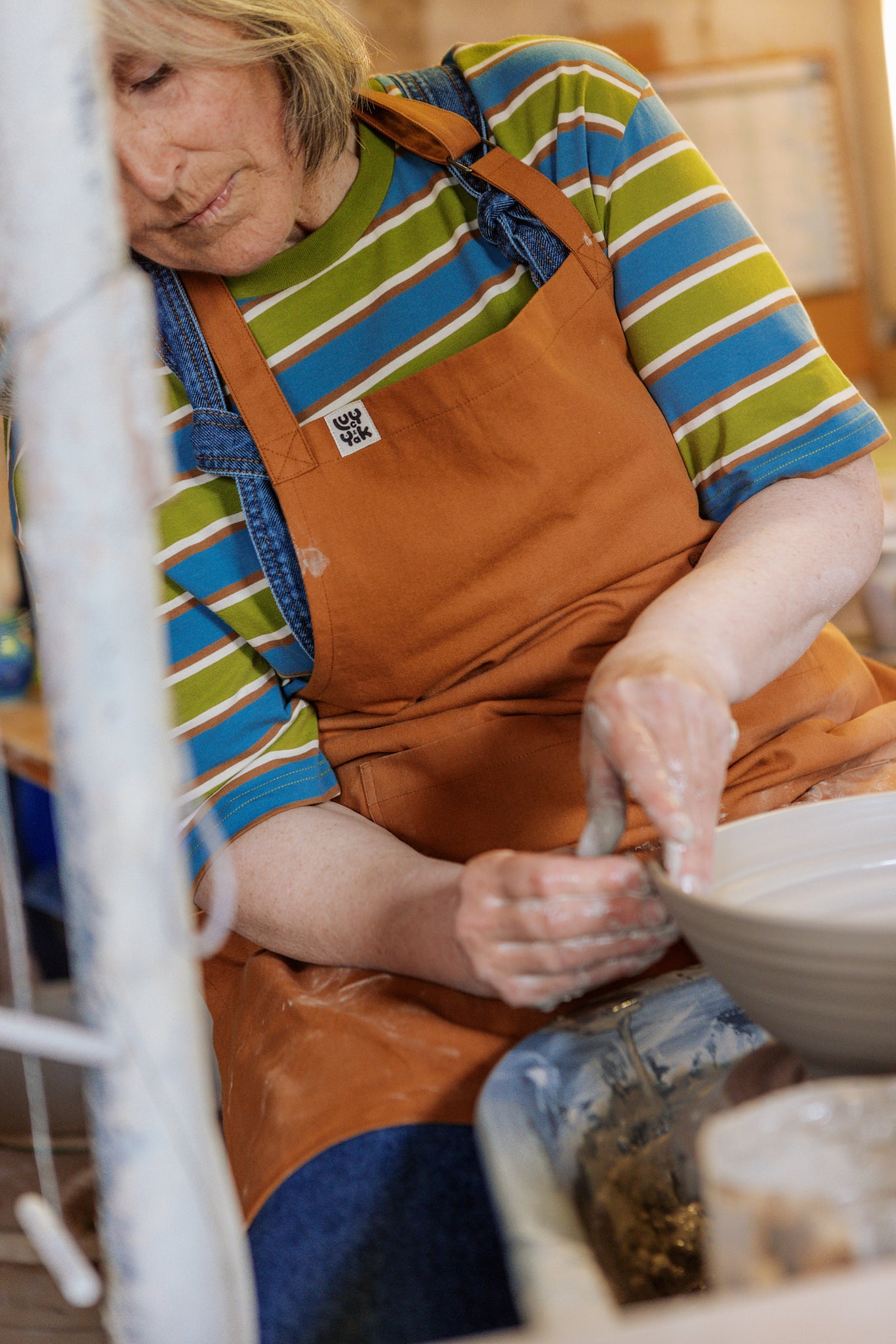 Person in a striped Lucy & Yak shirt and brown Lucy & Yak apron working with clay in a pottery studio.